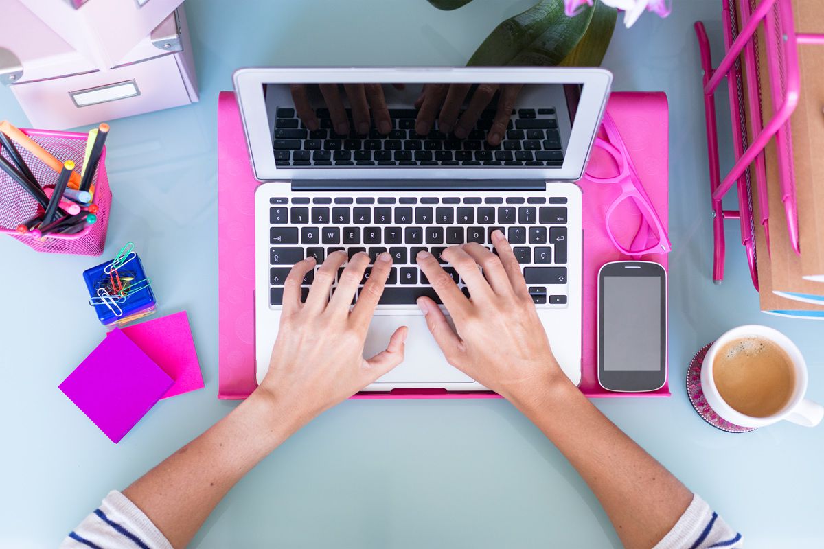 Schreibtischszene von oben aufgenommen: MacBook mit Händen auf der Tastatur, und pinkfarbenen Utensilien auf dem Tisch.
